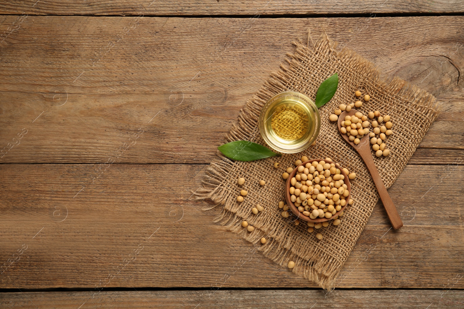 Photo of Glass bowl with oil, leaves and soybeans on wooden table, flat lay. Space for text