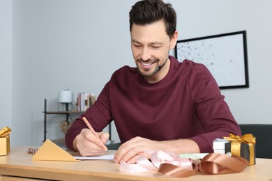 Happy man writing message in greeting card at wooden table in room