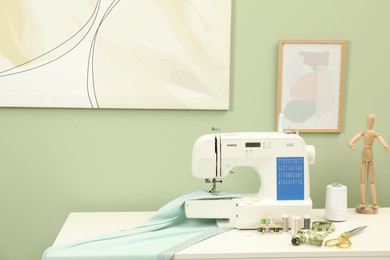 Modern sewing machine with cloth and craft accessories on white table near light green wall