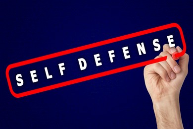 Image of Man contouring words Self Defense with marker glass board against blue background, closeup