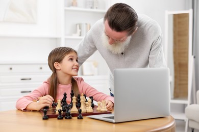 Photo of Grandfather teaching his granddaughter to play chess following online lesson at home