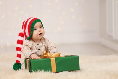 Photo of Cute baby in elf hat with Christmas gift on fluffy carpet against blurred festive lights, space for text. Winter holiday