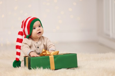 Cute baby in elf hat with Christmas gift on fluffy carpet against blurred festive lights, space for text. Winter holiday