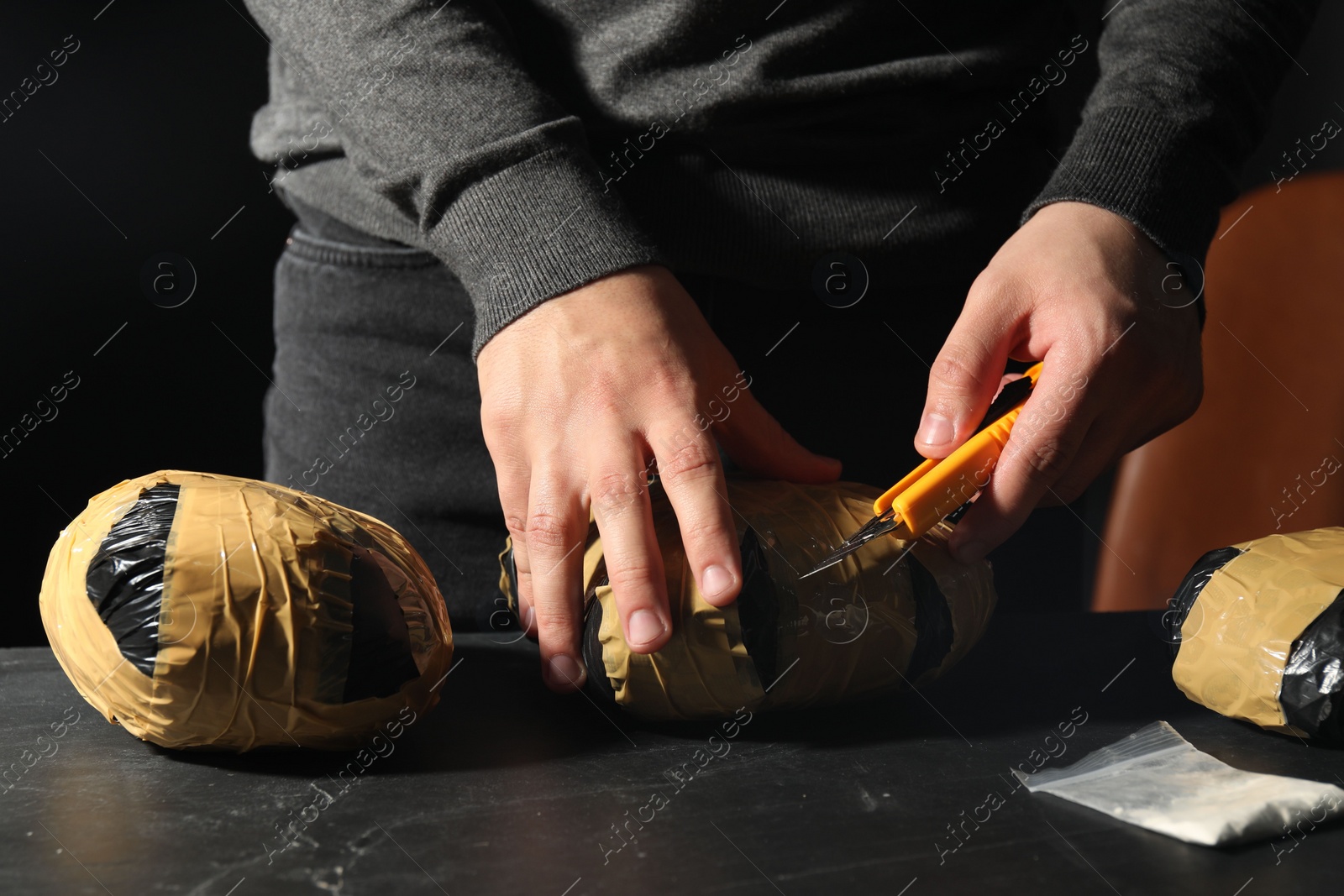 Photo of Smuggling and drug trafficking. Man opening package of narcotics with box cutter at dark table, closeup