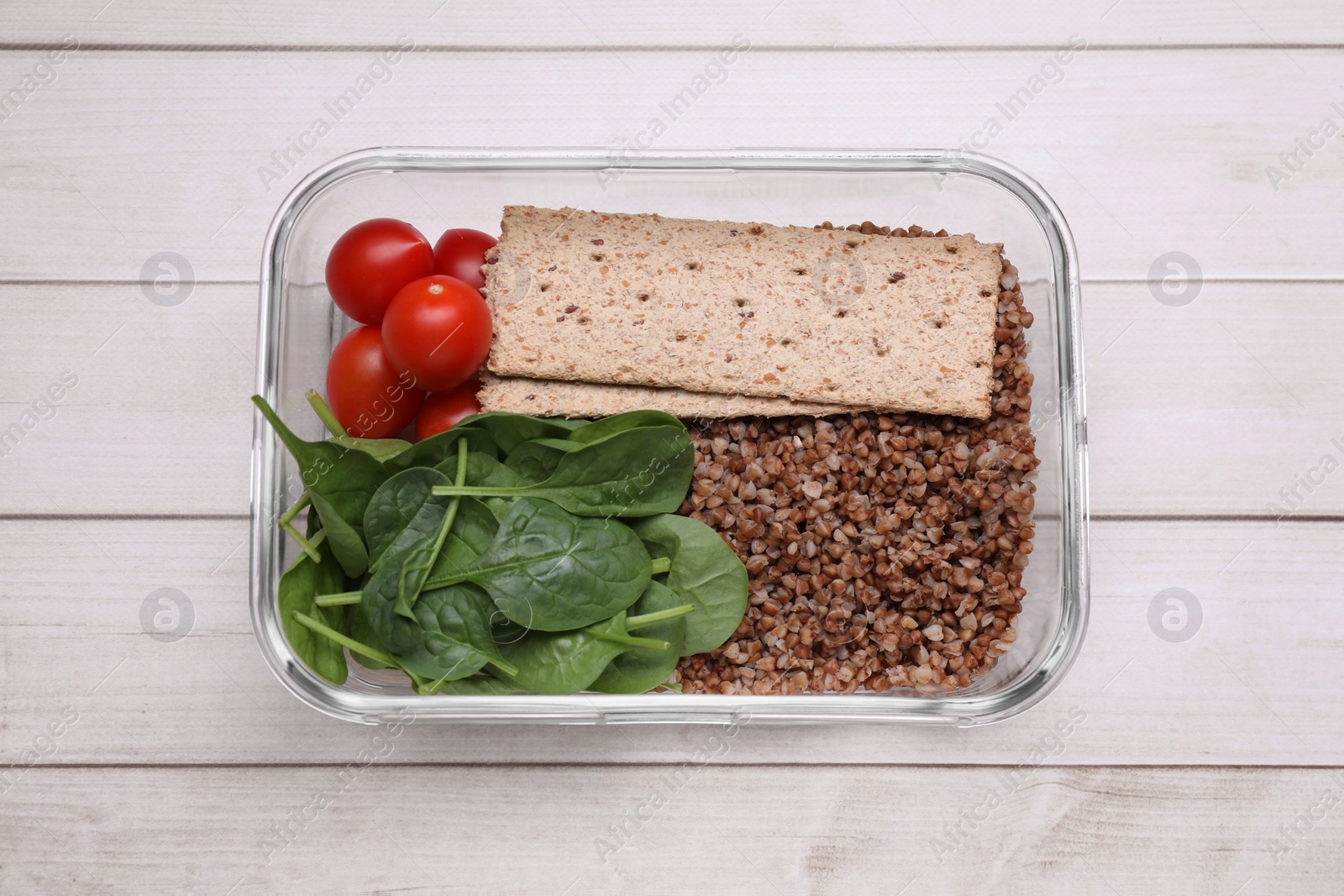 Photo of Glass container with buckwheat, fresh tomato, spinach and crispbreads on white wooden table, top view