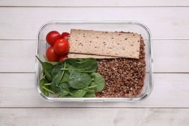 Photo of Glass container with buckwheat, fresh tomato, spinach and crispbreads on white wooden table, top view