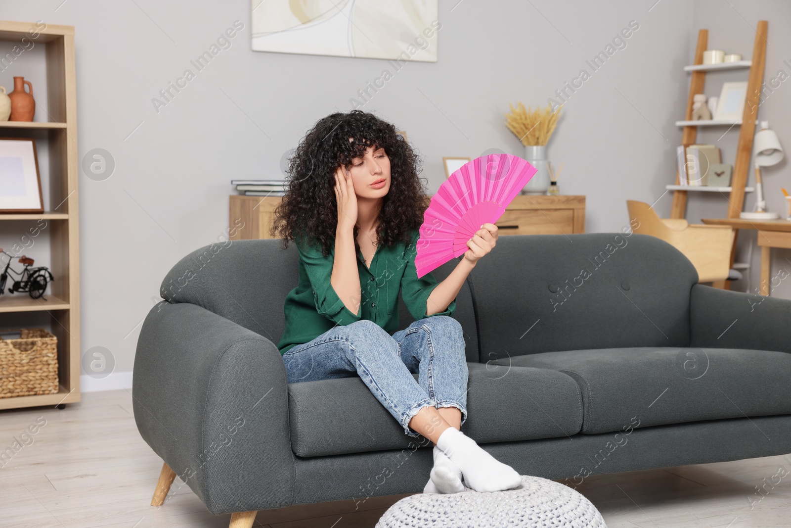 Photo of Young woman waving pink hand fan to cool herself on sofa at home