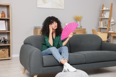 Young woman waving pink hand fan to cool herself on sofa at home