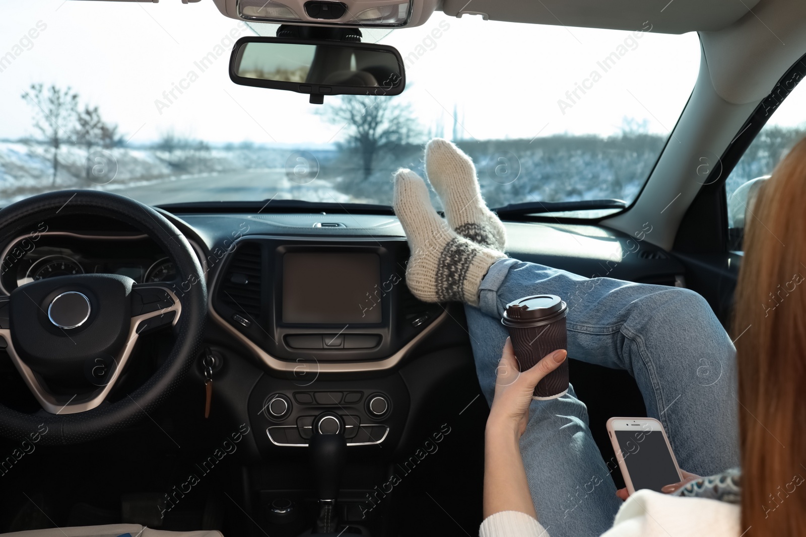 Photo of Young woman with phone in warm socks holding her legs on car dashboard and drinking coffee. Cozy atmosphere