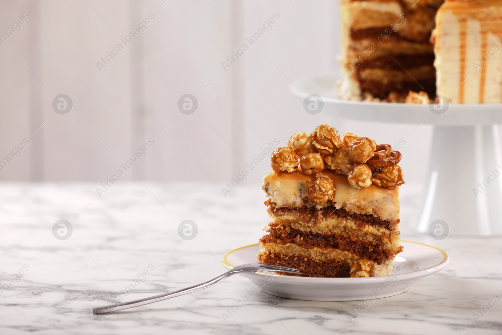 Photo of Piece of caramel drip cake decorated with popcorn and pretzels on white marble table, space for text