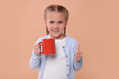 Happy girl with red ceramic mug showing thumbs up on beige background