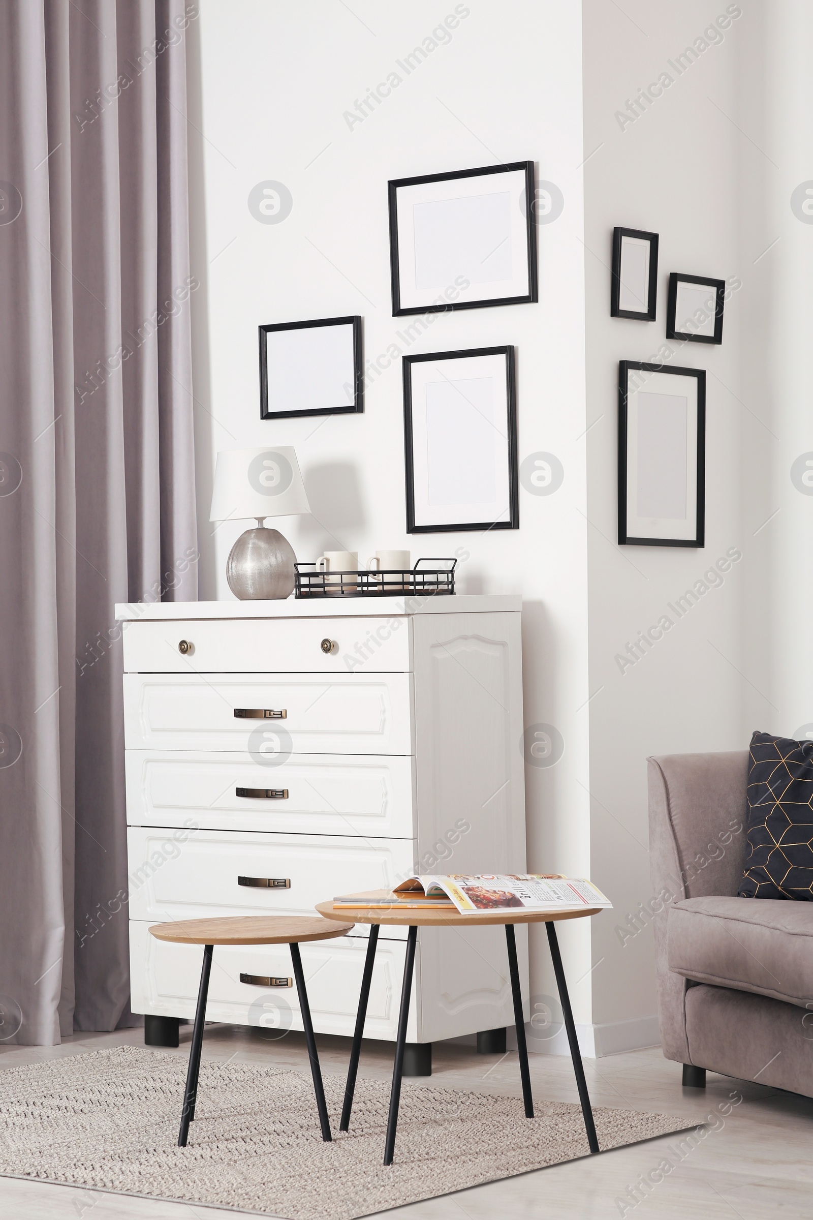 Photo of Empty frames hanging on white wall, chest of drawers and wooden tables indoors