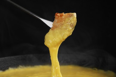 Dipping piece of bread into fondue pot with tasty melted cheese against dark gray background, closeup