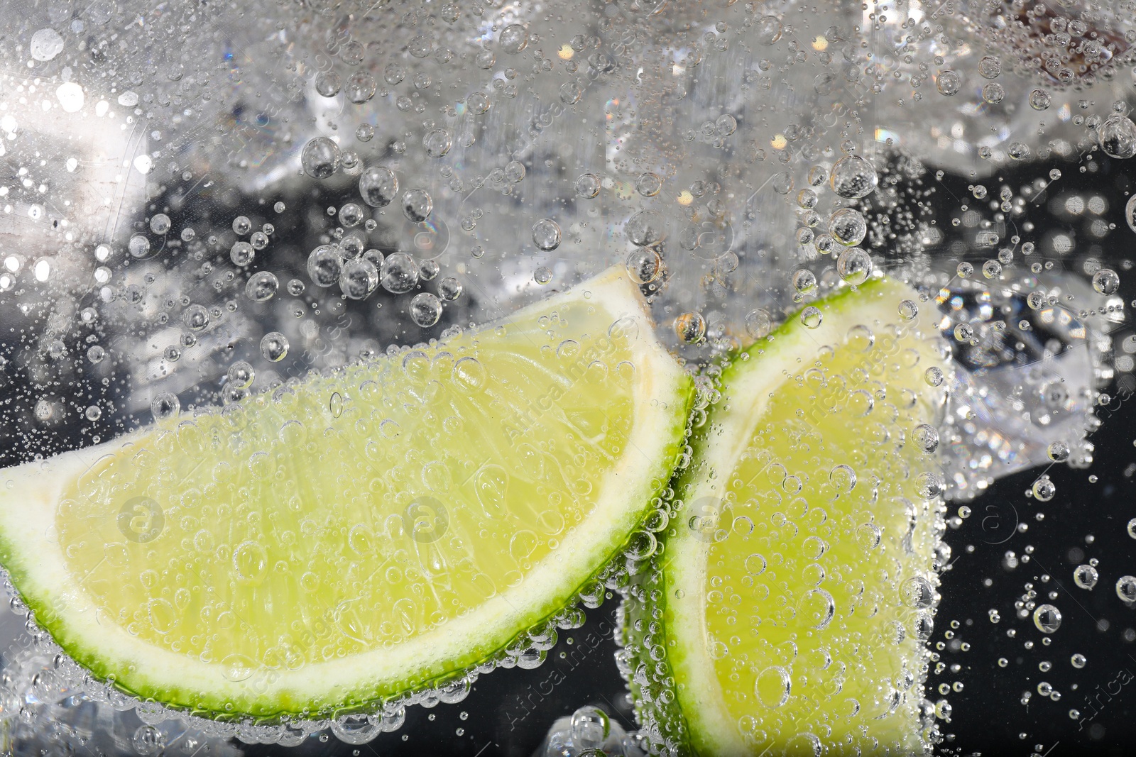 Photo of Juicy lime slices and ice cubes in soda water against black background, closeup