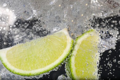 Juicy lime slices and ice cubes in soda water against black background, closeup