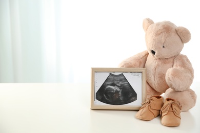 Ultrasound picture, teddy bear and baby shoes on table against light background. Space for text