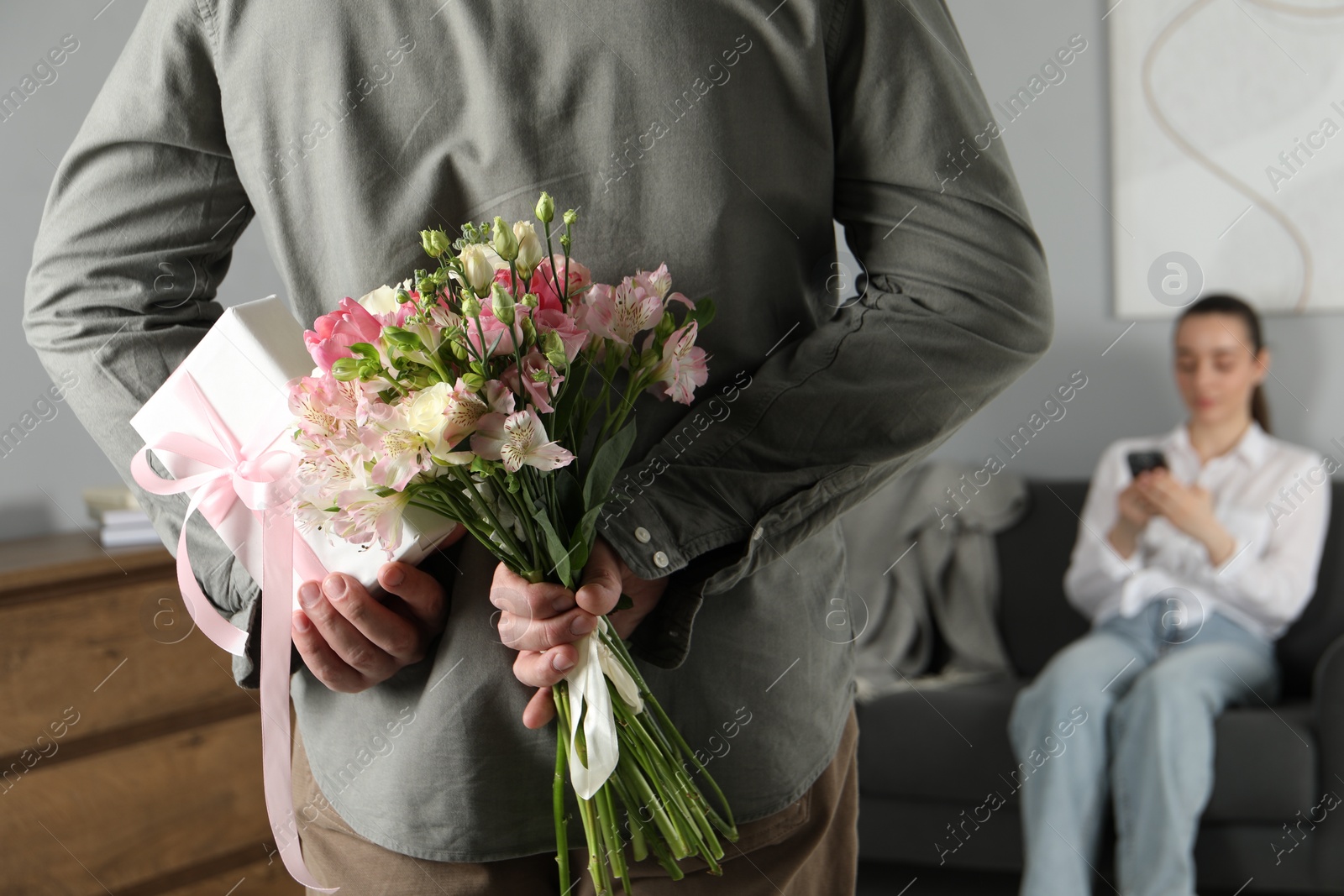 Photo of Man hiding bouquet of flowers and present for his beloved woman indoors, closeup