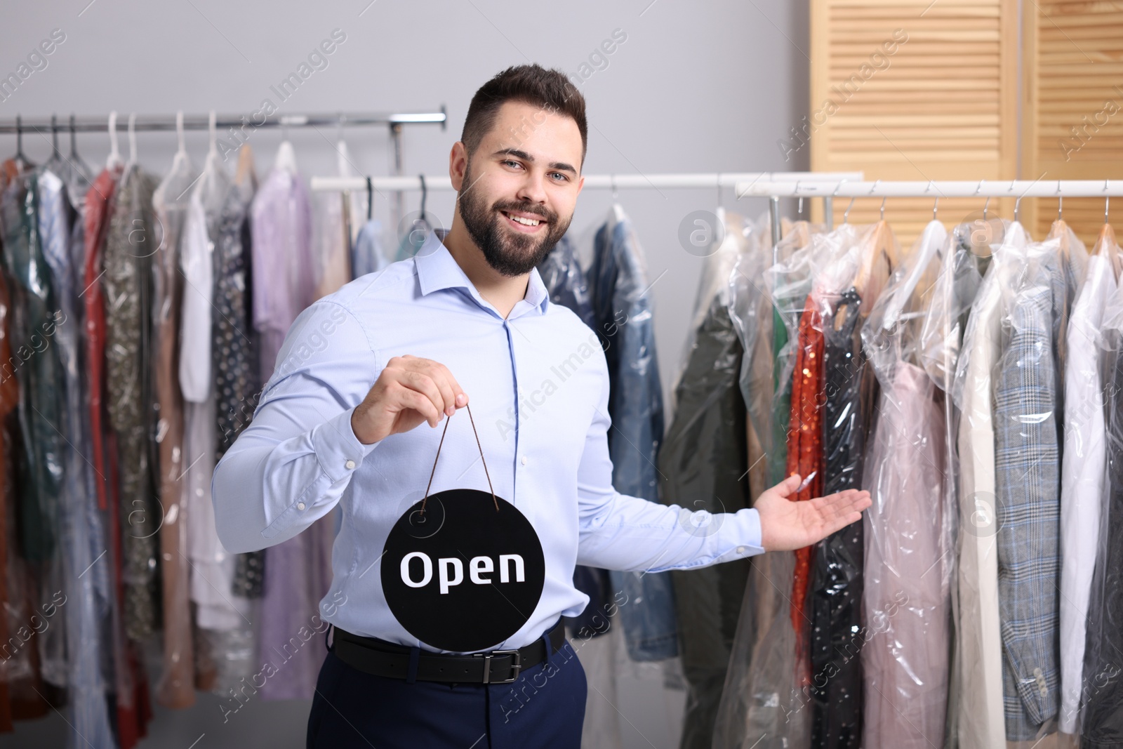 Photo of Dry-cleaning service. Happy worker holding Open sign near racks with clothes indoors