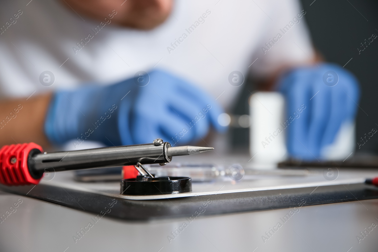 Photo of Soldering iron in focus and blurred technician repairing mobile phone at table, closeup with space for text