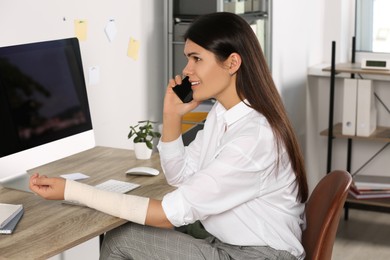 Young woman with arm wrapped in medical bandage talking on phone at workplace