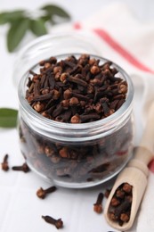 Photo of Aromatic cloves in glass jar and scoop on white table, closeup