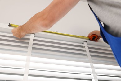 Worker in uniform using measuring tape while installing horizontal window blinds indoors, closeup