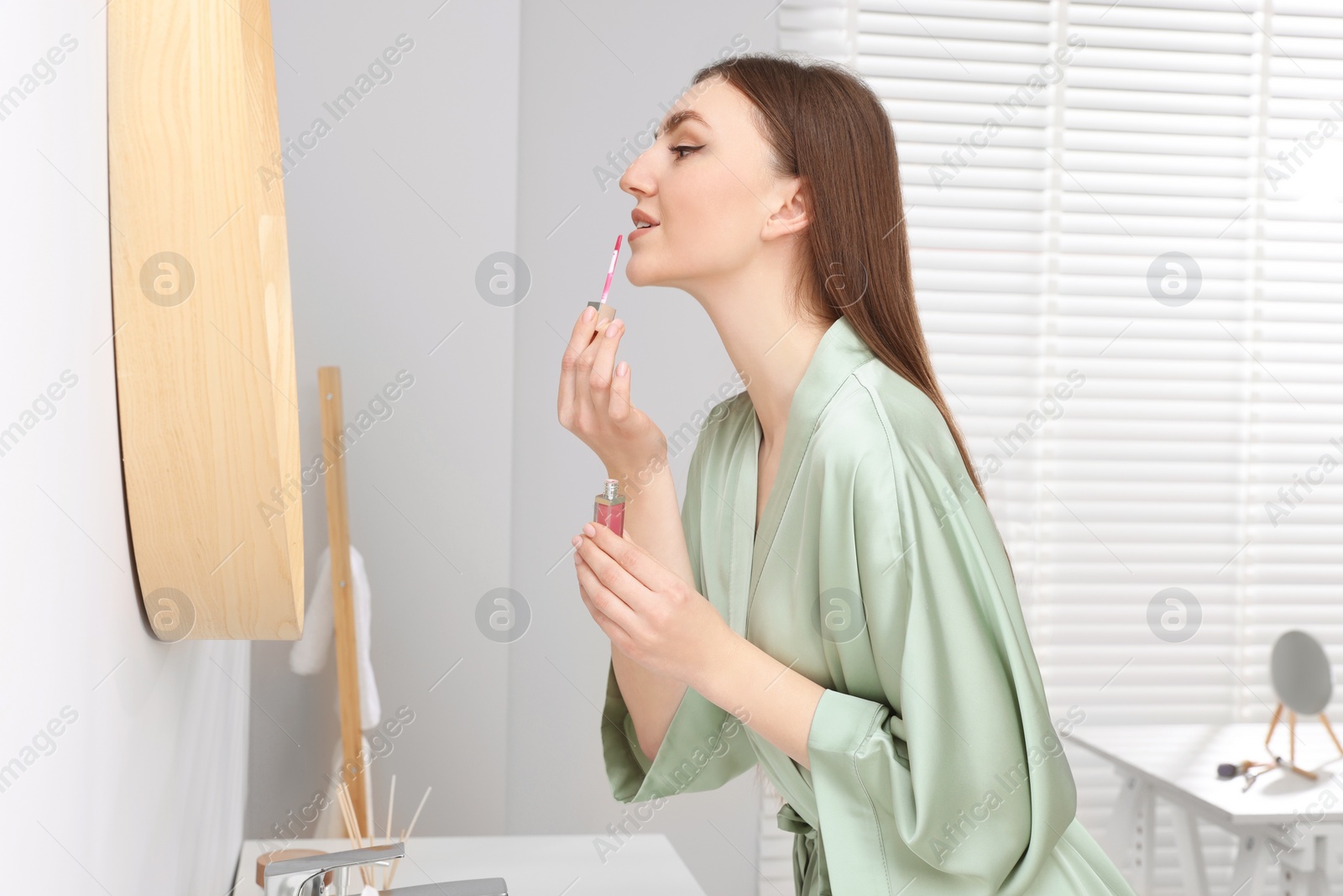 Photo of Beautiful young woman applying lipstick near mirror in bathroom