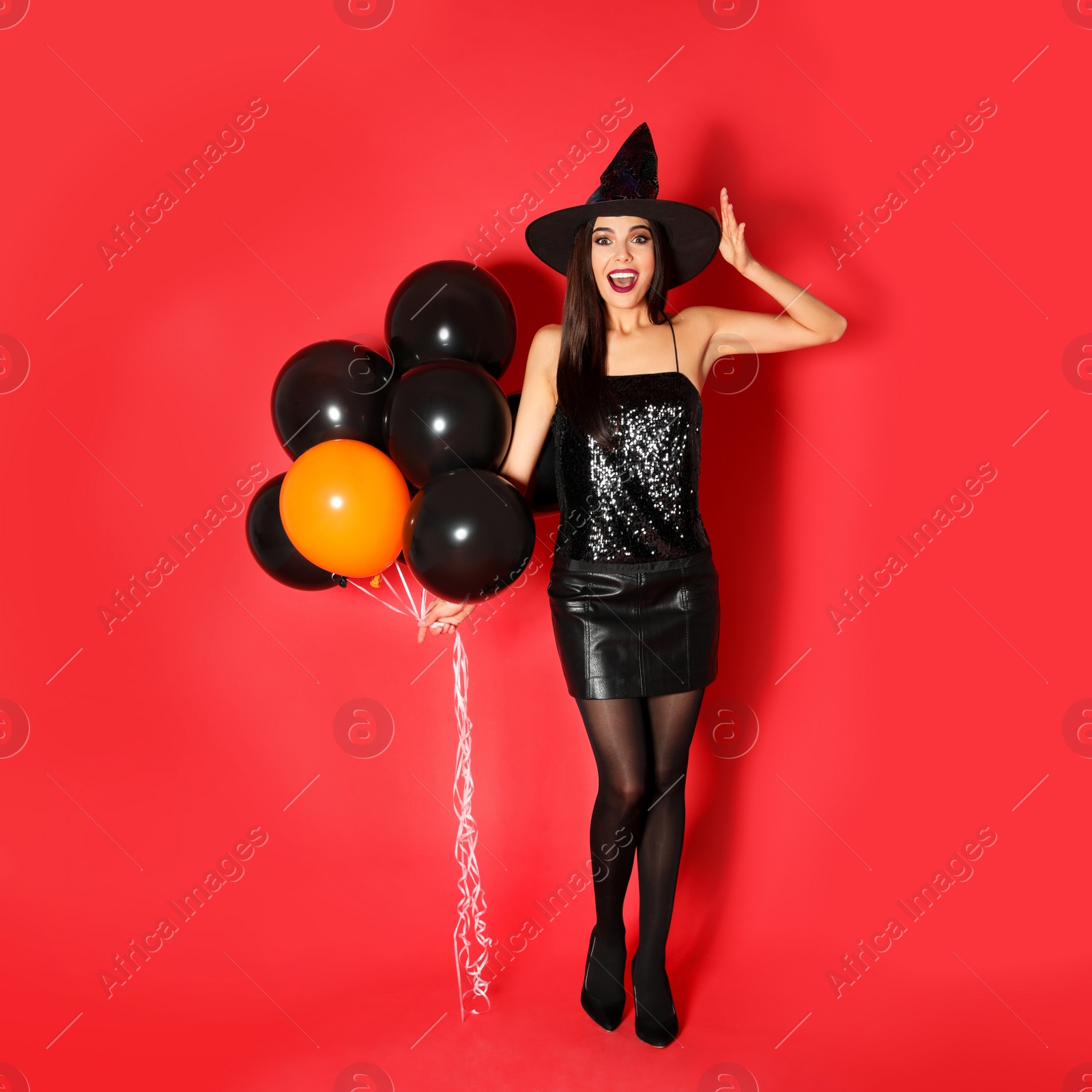 Photo of Beautiful young woman wearing witch costume with balloons for Halloween party on red background