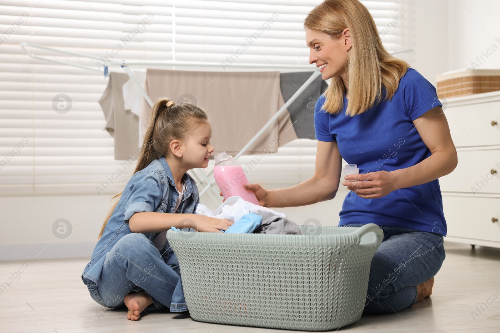 Photo of Mother and daughter sitting near basket with dirty clothes and smelling fabric softener in bathroom