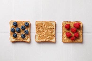 Photo of Delicious toasts with peanut butter, raspberries and blueberries on white tiled table, flat lay