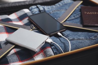 Photo of Smartphone charging with power bank, passport and clothes in open suitcase, closeup