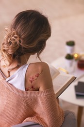 Image of Young woman with tattoo of beautiful sakura tree branch reading at home, back view