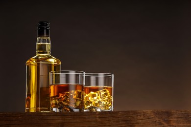 Whiskey with ice cubes in glasses and bottle on wooden table against brown background, space for text