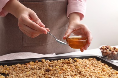 Photo of Making granola. Woman adding honey onto baking tray with mixture of oat flakes and other ingredients at white table in kitchen, closeup