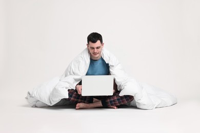 Happy man in pyjama wrapped in blanket using laptop on light grey background