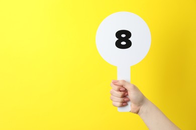 Photo of Woman holding auction paddle with number 8 on yellow background, closeup. Space for text