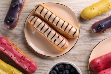 Different tasty glazed eclairs and blueberries on light wooden table, flat lay