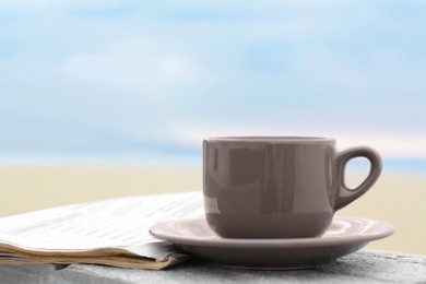 Photo of Ceramic cup of hot drink and newspaper on stone surface near sea in morning