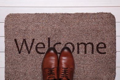 Door mat with word WELCOME and shoes on white wooden floor, top view