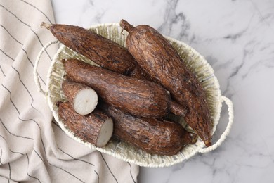 Whole and cut cassava roots in wicker basket on white marble table, top view