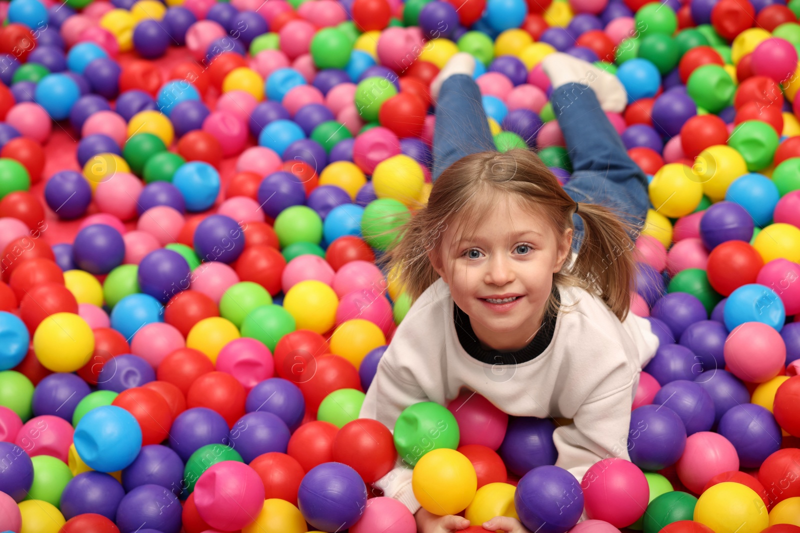 Photo of Happy little girl lying on colorful balls in ball pit, space for text