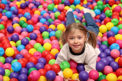 Photo of Happy little girl lying on colorful balls in ball pit, space for text
