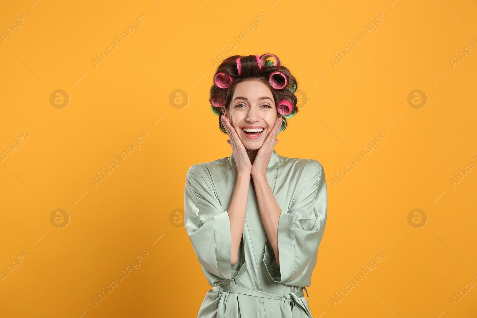 Photo of Happy young woman in silk bathrobe with hair curlers on yellow background