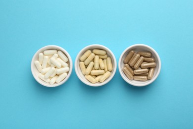 Different vitamin capsules in bowls on light blue background, flat lay