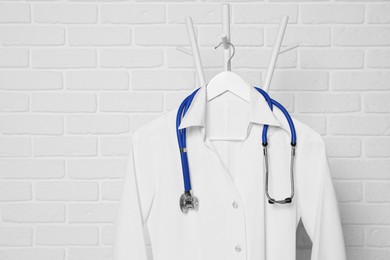 Medical uniform and stethoscope hanging on rack near white brick wall. Space for text