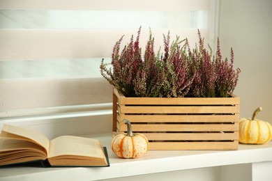 Beautiful heather flowers in crate, book and pumpkins on white windowsill indoors