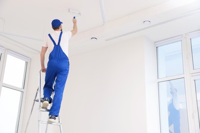 Worker in uniform painting ceiling with roller on stepladder indoors, back view. Space for text