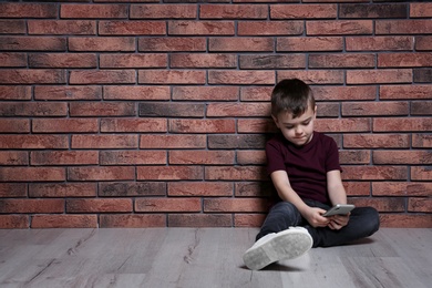 Photo of Sad little boy with mobile phone sitting on floor near brick wall. Space for text