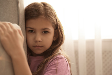 Photo of Sad little girl hiding behind sofa at home. Domestic violence concept