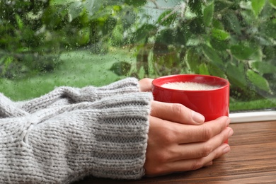 Photo of Woman with cup of coffee near window on rainy day, closeup